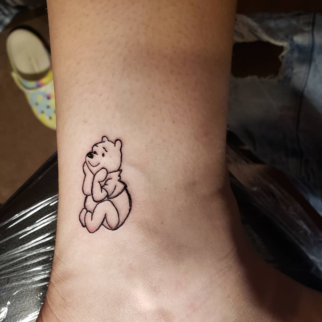 Small outline tattoo of Winnie the Pooh sitting with a thoughtful expression on a person's ankle